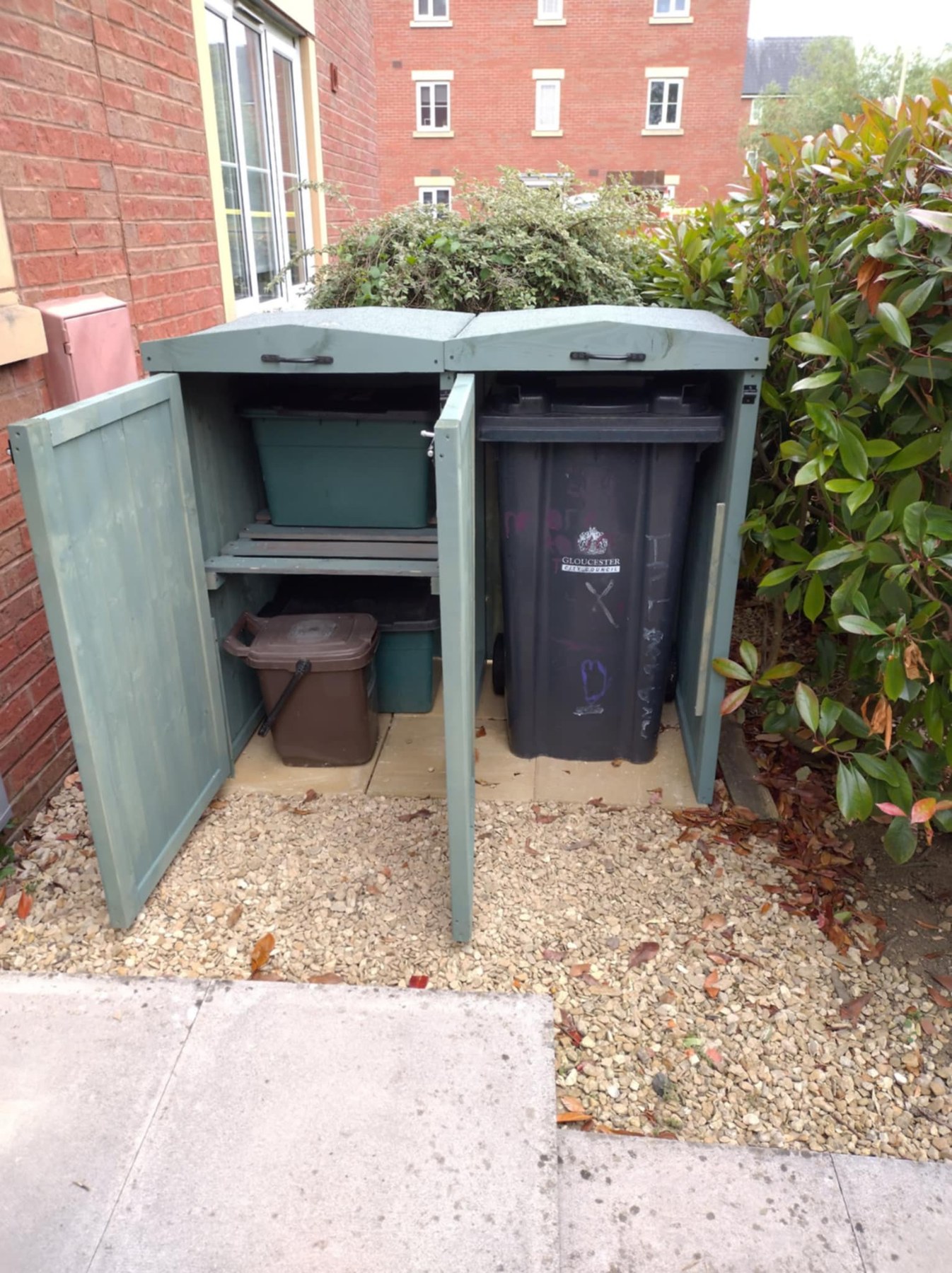 Open sage green wooden bin store showing a wheelie bin compartment and shelves for recycling containers